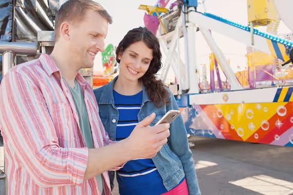 Pasangan turis dewasa yang bersemangat mengunjungi taman hiburan menggunakan ponsel pintar, berjejaring dan berbagi, tersenyum bersama di luar ruangan yang cerah. Shutterstock/MJTH